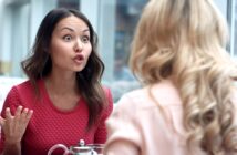 A woman wearing a red shirt is sitting at a table, gesturing with her hand and appearing to be in an intense conversation with another woman who has blonde, curly hair and is wearing a light pink shirt. They are both indoors near large windows.