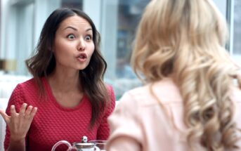 A woman wearing a red shirt is sitting at a table, gesturing with her hand and appearing to be in an intense conversation with another woman who has blonde, curly hair and is wearing a light pink shirt. They are both indoors near large windows.