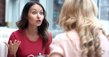 A woman wearing a red shirt is sitting at a table, gesturing with her hand and appearing to be in an intense conversation with another woman who has blonde, curly hair and is wearing a light pink shirt. They are both indoors near large windows.