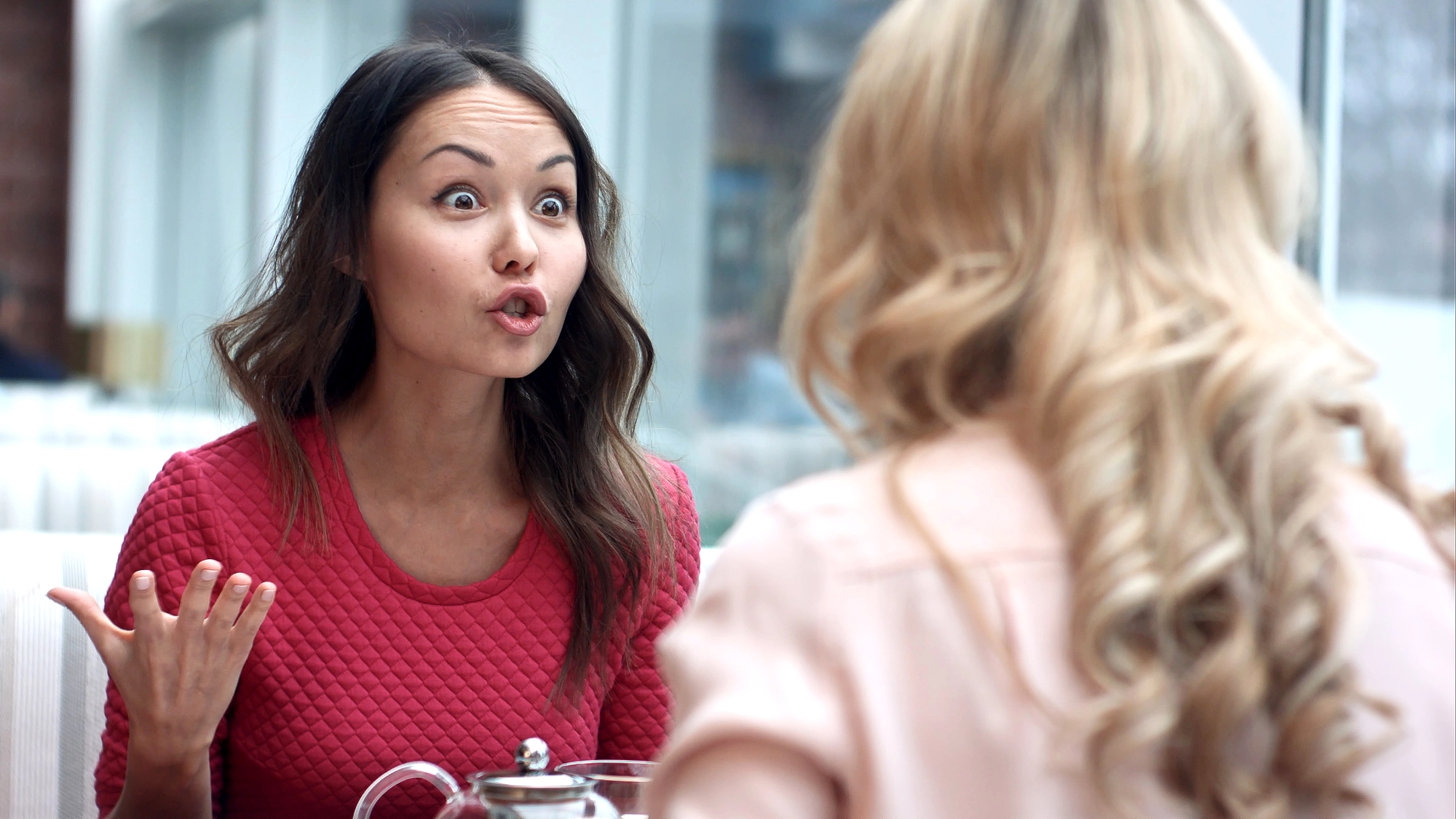 A woman wearing a red shirt is sitting at a table, gesturing with her hand and appearing to be in an intense conversation with another woman who has blonde, curly hair and is wearing a light pink shirt. They are both indoors near large windows.