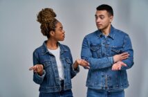 A woman and a man are standing and talking, both wearing denim jackets. The woman has braided hair and gestures with her palms up. The man has short hair, crossing his arms, and appears to be responding. They are on a neutral background.