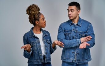 A woman and a man are standing and talking, both wearing denim jackets. The woman has braided hair and gestures with her palms up. The man has short hair, crossing his arms, and appears to be responding. They are on a neutral background.