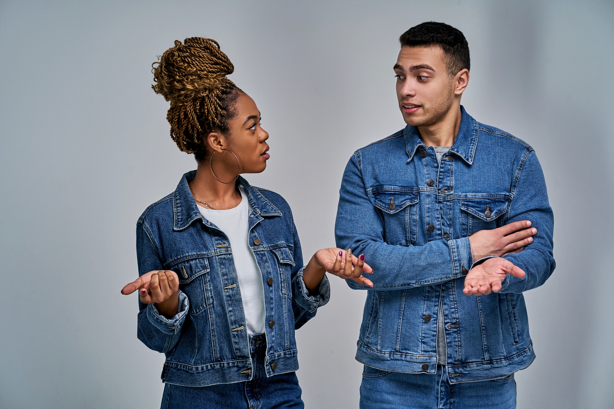 A woman and a man are standing and talking, both wearing denim jackets. The woman has braided hair and gestures with her palms up. The man has short hair, crossing his arms, and appears to be responding. They are on a neutral background.