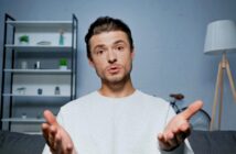 A person with short dark hair is sitting on a couch with hands gesturing towards the camera. In the background, there's a bookshelf with plants and decor, a bicycle, and a floor lamp. The person appears to be speaking or explaining something.