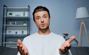 A person with short dark hair is sitting on a couch with hands gesturing towards the camera. In the background, there's a bookshelf with plants and decor, a bicycle, and a floor lamp. The person appears to be speaking or explaining something.