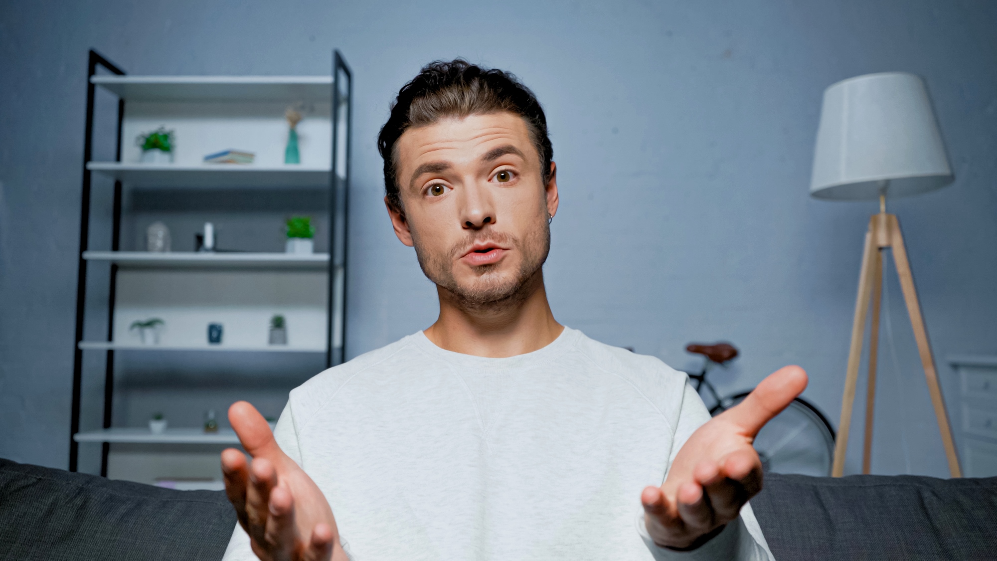 A person with short dark hair is sitting on a couch with hands gesturing towards the camera. In the background, there's a bookshelf with plants and decor, a bicycle, and a floor lamp. The person appears to be speaking or explaining something.