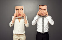 Two people standing against a gray background hold photos of faces over their own. The person on the left has a skeptical female face, while the person on the right holds a suspicious male face. Both wear formal white tops.