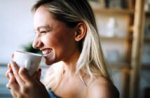 A woman with long blonde hair smiles joyfully while holding a white mug. She is indoors, and the background is softly blurred, suggesting a cozy setting. Her eyes are closed, conveying a sense of contentment and warmth.