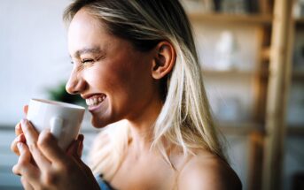 A woman with long blonde hair smiles joyfully while holding a white mug. She is indoors, and the background is softly blurred, suggesting a cozy setting. Her eyes are closed, conveying a sense of contentment and warmth.