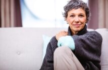 A person with short gray hair sits on a light-colored couch, wearing a dark sweater. They are smiling gently, resting one arm on their knee. The background is softly blurred, creating a cozy atmosphere.