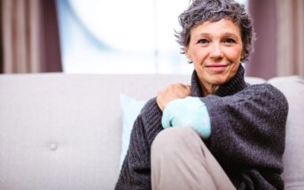 A person with short gray hair sits on a light-colored couch, wearing a dark sweater. They are smiling gently, resting one arm on their knee. The background is softly blurred, creating a cozy atmosphere.