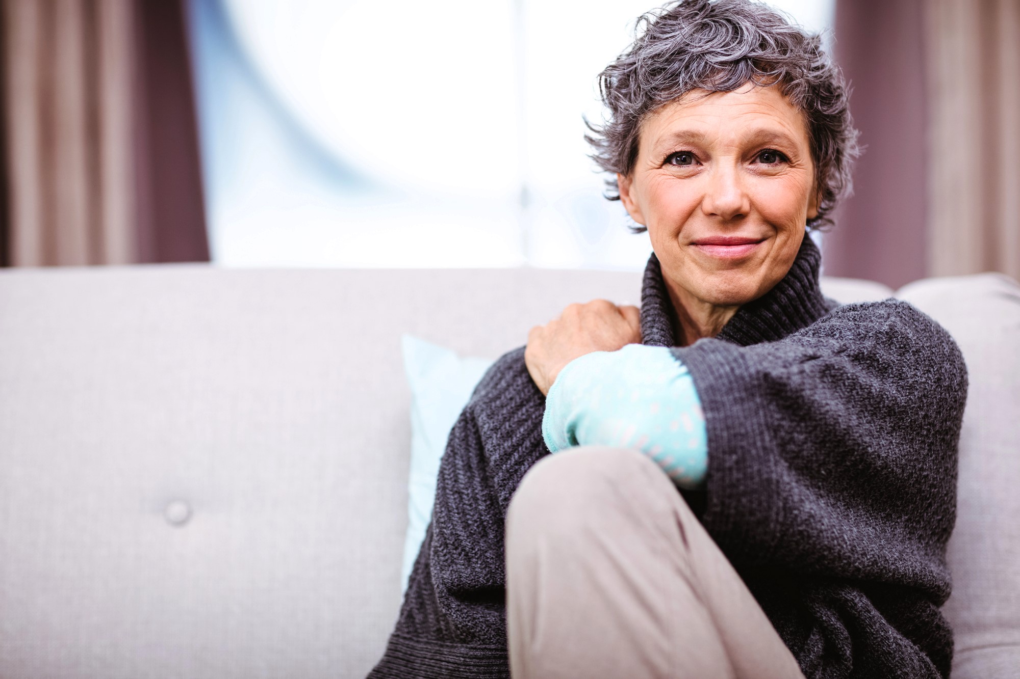 A person with short gray hair sits on a light-colored couch, wearing a dark sweater. They are smiling gently, resting one arm on their knee. The background is softly blurred, creating a cozy atmosphere.