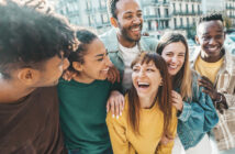 A diverse group of friends stands closely together, smiling and laughing in an urban setting. The background shows buildings and a lively street. The mood is joyful and warm, capturing a moment of friendship and camaraderie.