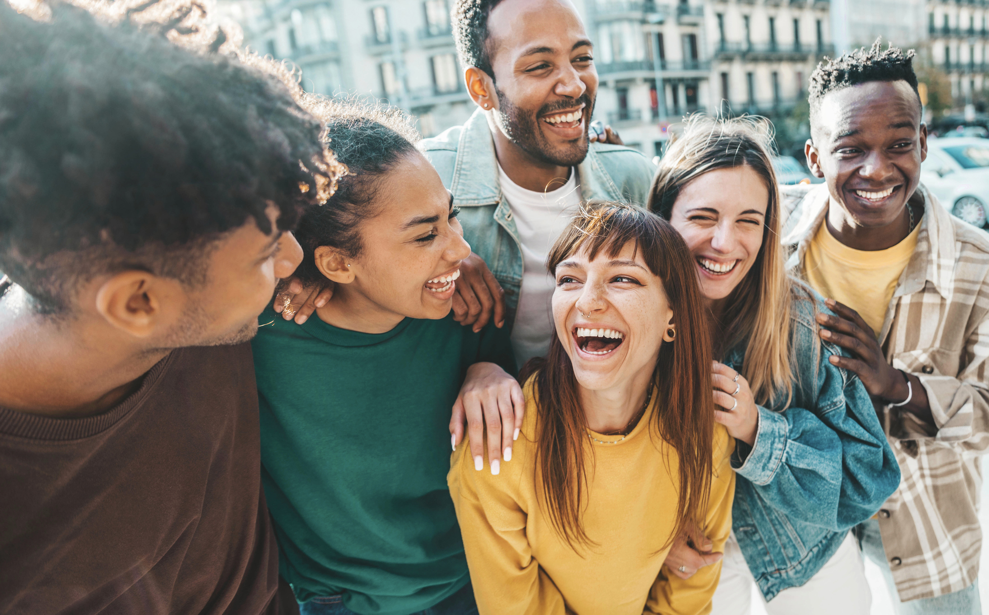 A diverse group of friends stands closely together, smiling and laughing in an urban setting. The background shows buildings and a lively street. The mood is joyful and warm, capturing a moment of friendship and camaraderie.