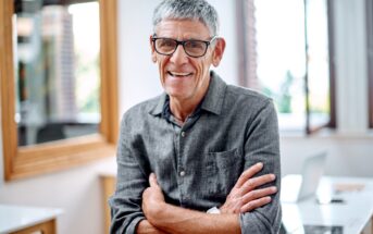 An older man with short gray hair and glasses smiles, standing with arms crossed in a bright office setting. He's wearing a gray button-up shirt, and a laptop is visible on the desk behind him.