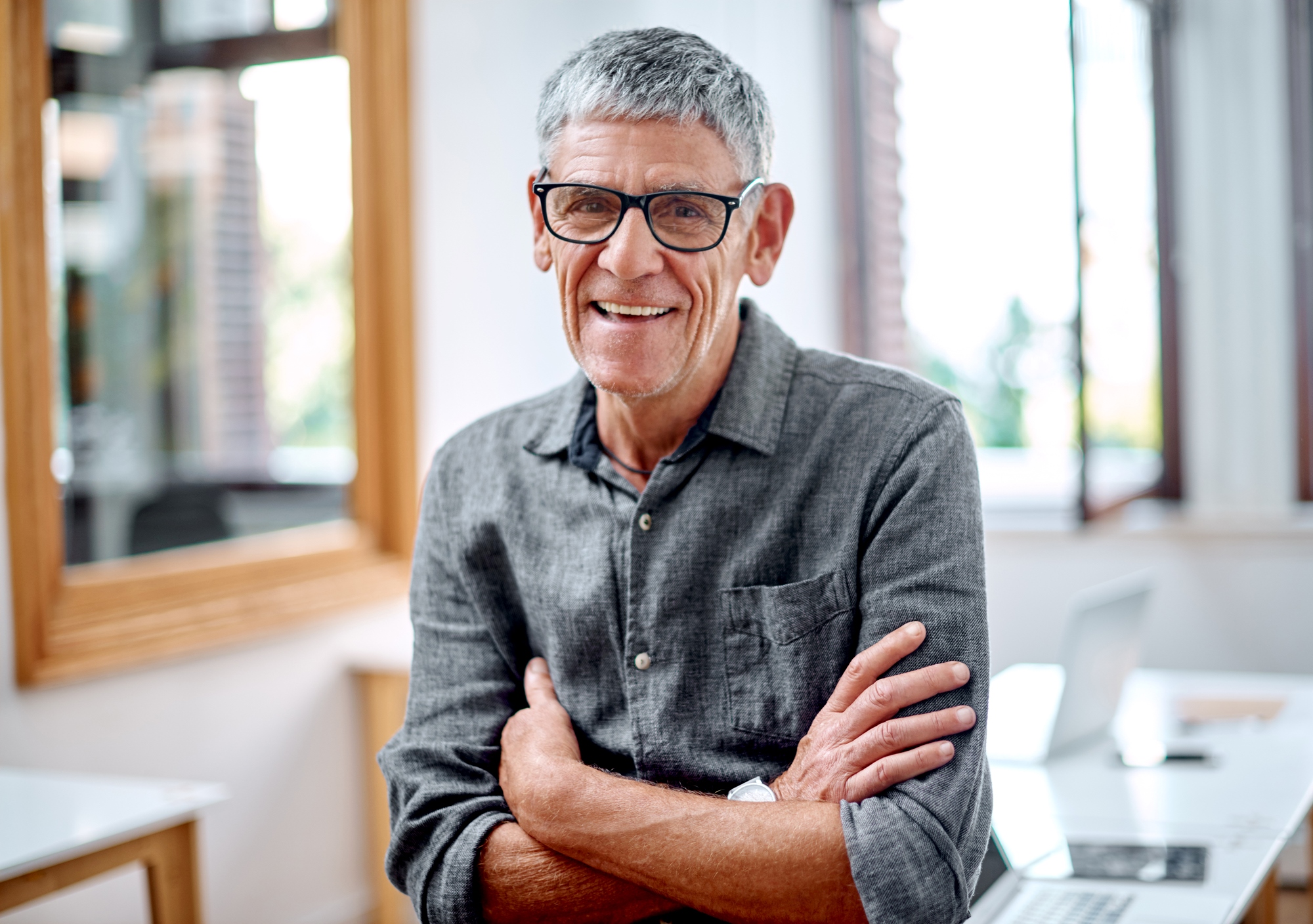 An older man with short gray hair and glasses smiles, standing with arms crossed in a bright office setting. He's wearing a gray button-up shirt, and a laptop is visible on the desk behind him.