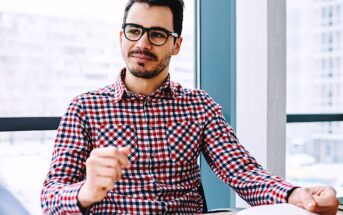 A man wearing glasses and a red and blue checkered shirt sits at a desk, holding a pen in his hand. He appears thoughtful and is looking slightly to the side. The background shows large windows with a blurred cityscape view.