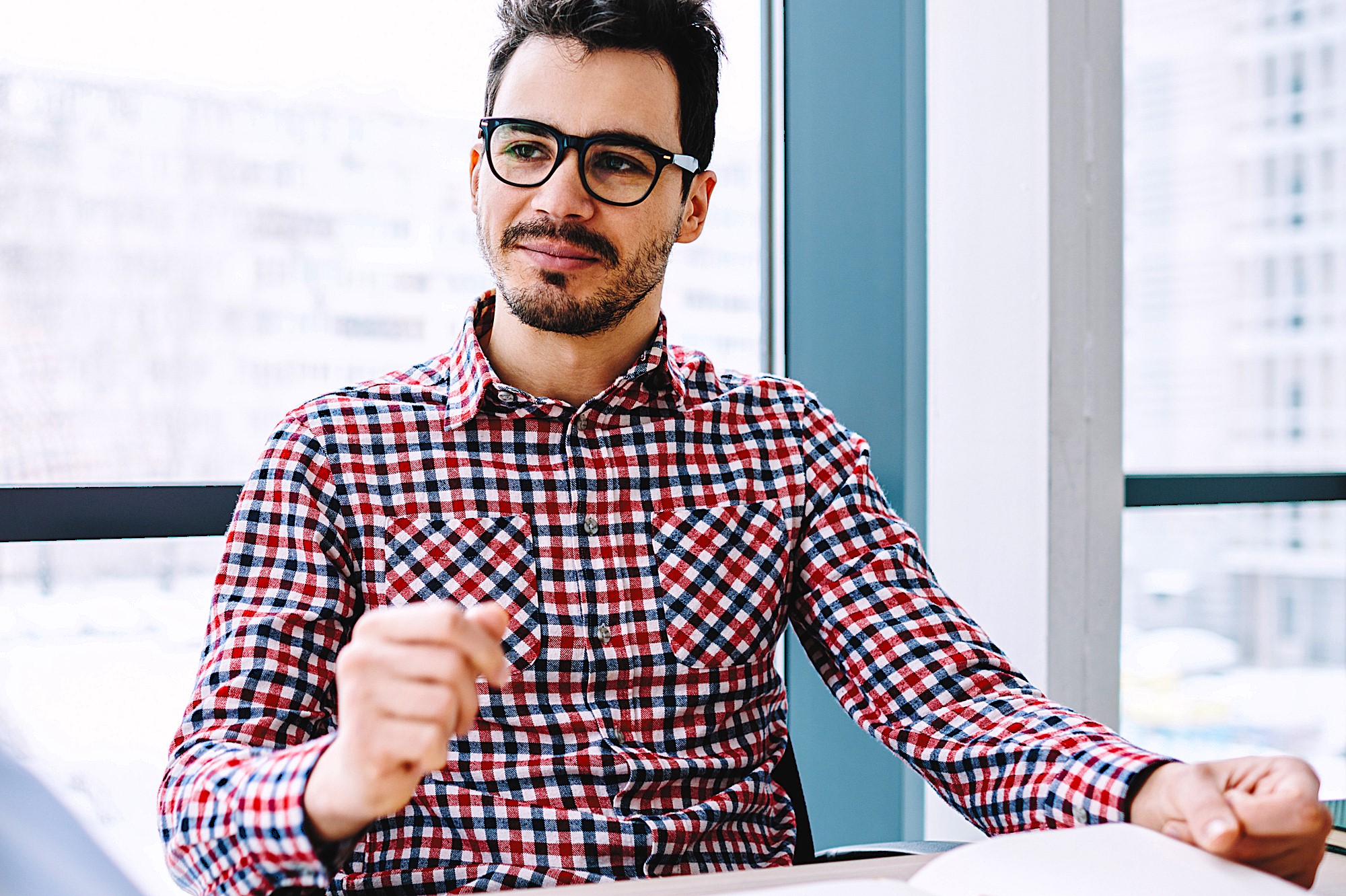 A man wearing glasses and a red and blue checkered shirt sits at a desk, holding a pen in his hand. He appears thoughtful and is looking slightly to the side. The background shows large windows with a blurred cityscape view.