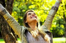 A woman outdoors with long hair and a light jacket smiles joyfully. She has her arms raised towards the sky, standing in front of a tree with green foliage, enjoying a sunny day.