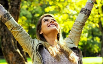 A woman outdoors with long hair and a light jacket smiles joyfully. She has her arms raised towards the sky, standing in front of a tree with green foliage, enjoying a sunny day.