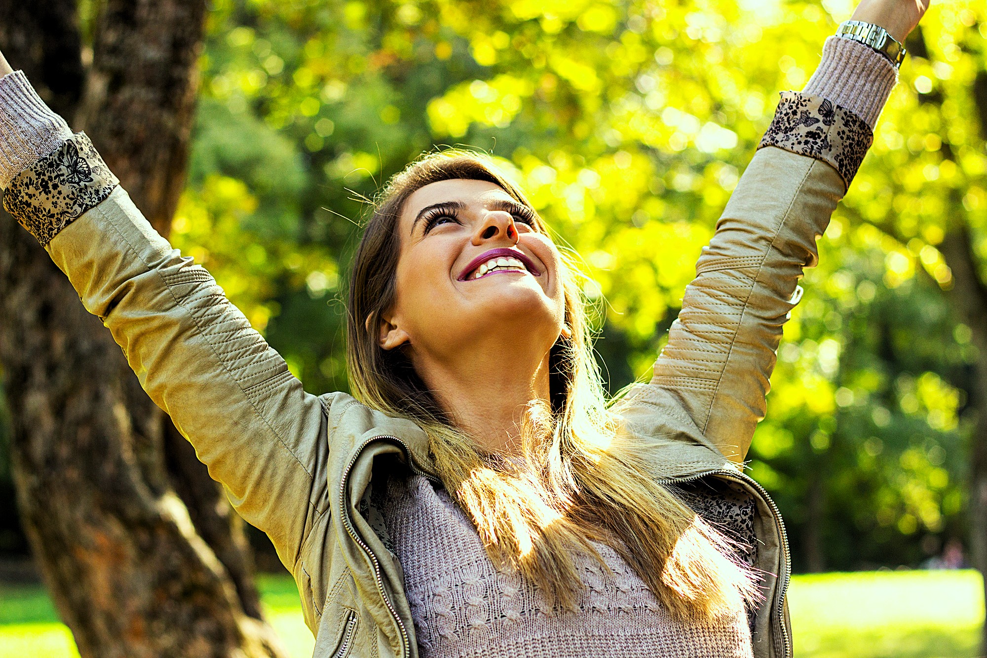 A woman outdoors with long hair and a light jacket smiles joyfully. She has her arms raised towards the sky, standing in front of a tree with green foliage, enjoying a sunny day.