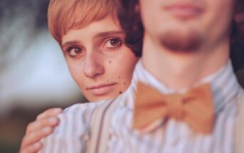 A close-up of a person with short hair embracing another person from behind. The foreground person wears a striped shirt and a bow tie, while the background person gazes softly at the camera. The atmosphere is warm and intimate.