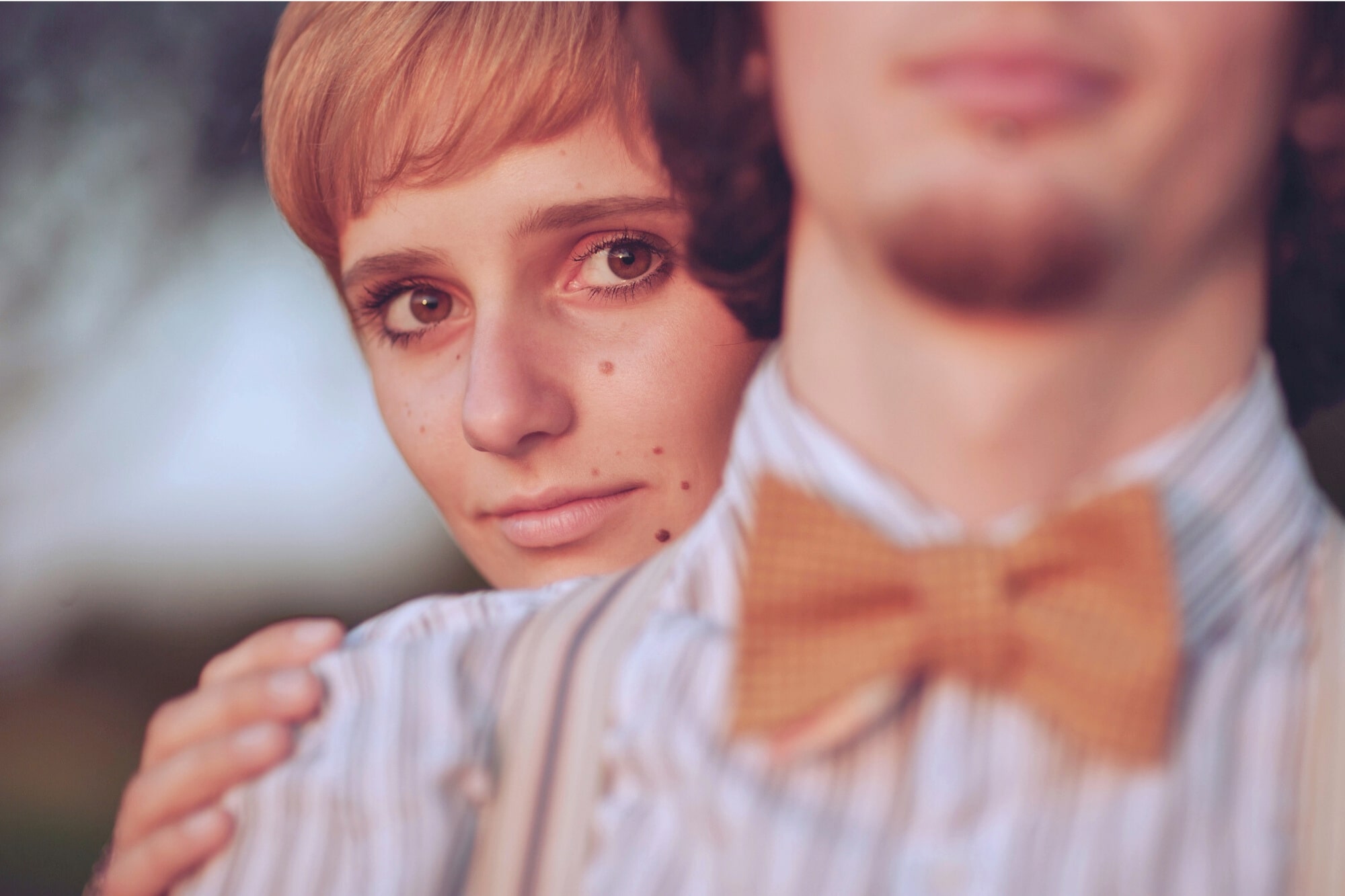 A close-up of a person with short hair embracing another person from behind. The foreground person wears a striped shirt and a bow tie, while the background person gazes softly at the camera. The atmosphere is warm and intimate.