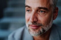 A middle-aged man with short gray hair and a beard gazes thoughtfully to the side. He is wearing a gray jacket, and the background is blurred, suggesting an outdoor setting. The image captures a calm and introspective moment.