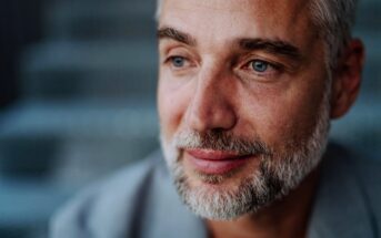 A middle-aged man with short gray hair and a beard gazes thoughtfully to the side. He is wearing a gray jacket, and the background is blurred, suggesting an outdoor setting. The image captures a calm and introspective moment.