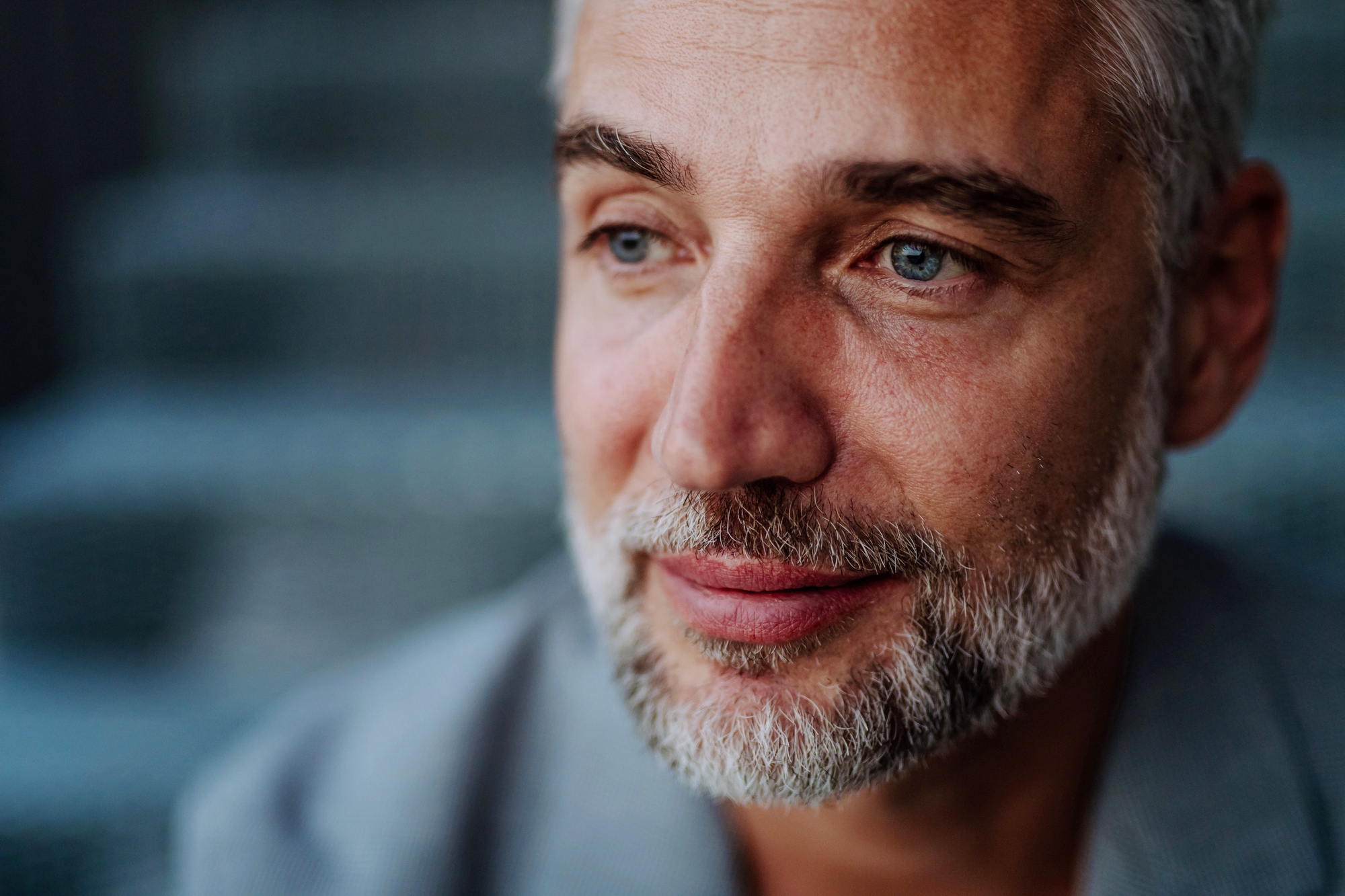 A middle-aged man with short gray hair and a beard gazes thoughtfully to the side. He is wearing a gray jacket, and the background is blurred, suggesting an outdoor setting. The image captures a calm and introspective moment.