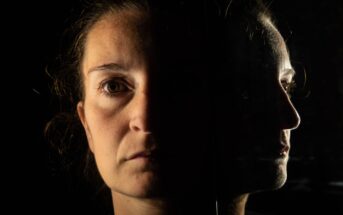 A woman's face is half-lit against a dark background, with her reflection visible in a mirror beside her. The lighting highlights her solemn expression, emphasizing the contrast between light and shadow.