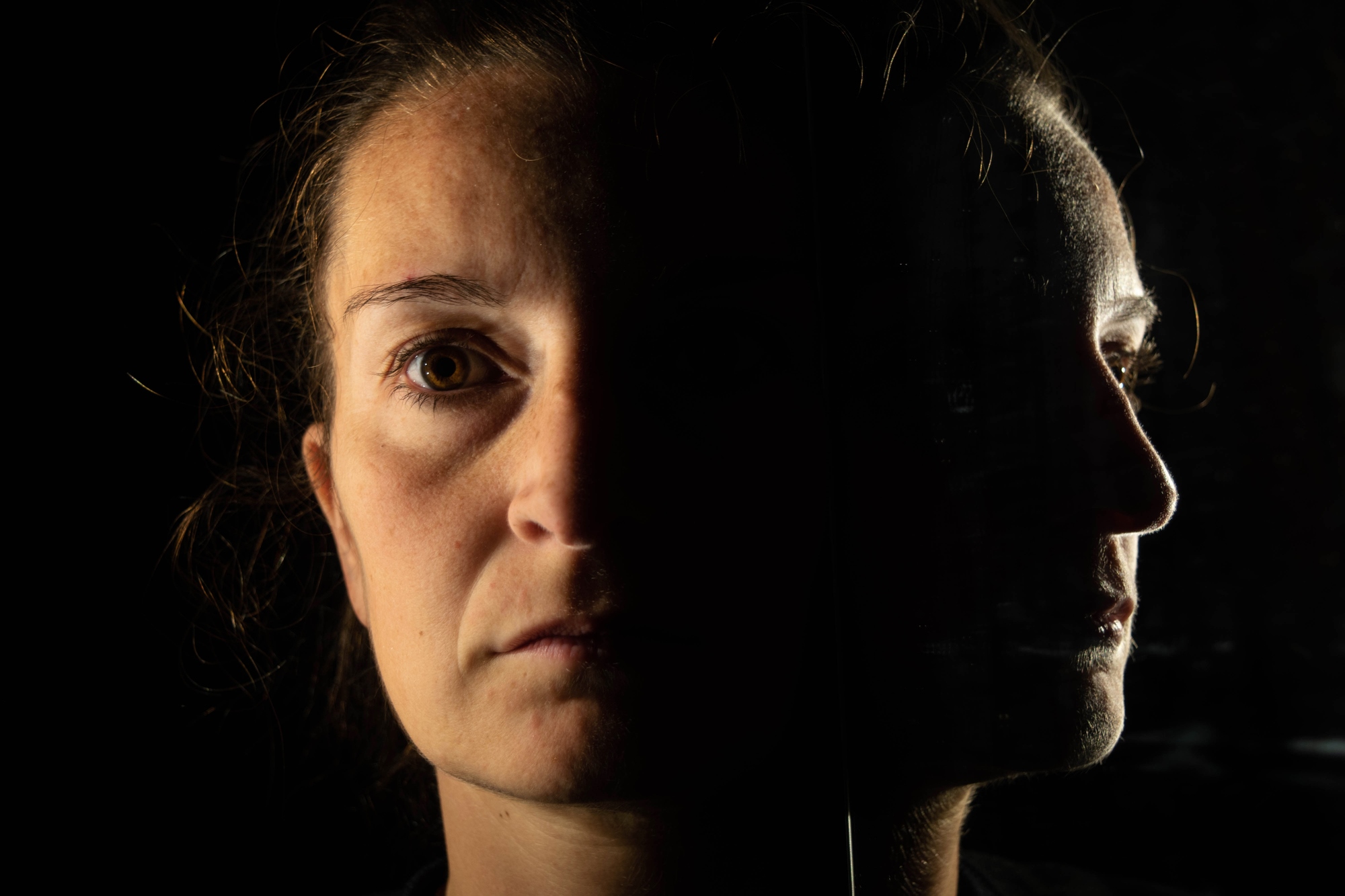 A woman's face is half-lit against a dark background, with her reflection visible in a mirror beside her. The lighting highlights her solemn expression, emphasizing the contrast between light and shadow.