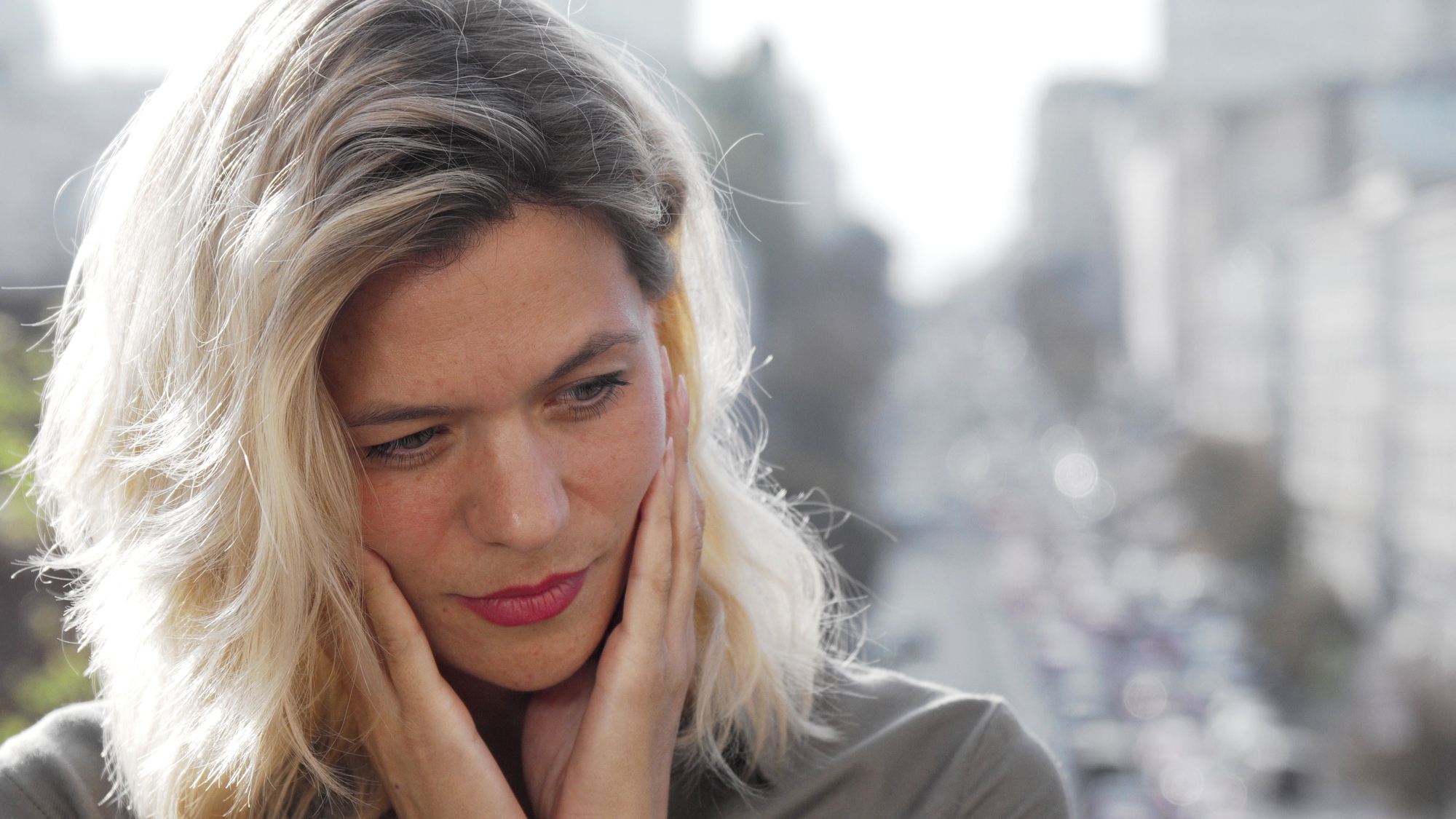 A woman with blonde hair holds her hands to her cheeks, gazing downwards. She appears thoughtful or concerned, standing outdoors with a blurred urban background.