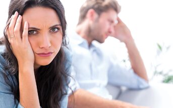 A woman with dark hair looks frustrated, resting her head on her hand, while a man in the background appears upset, sitting on a couch. Both wear casual clothing, and the background is bright and softly focused.