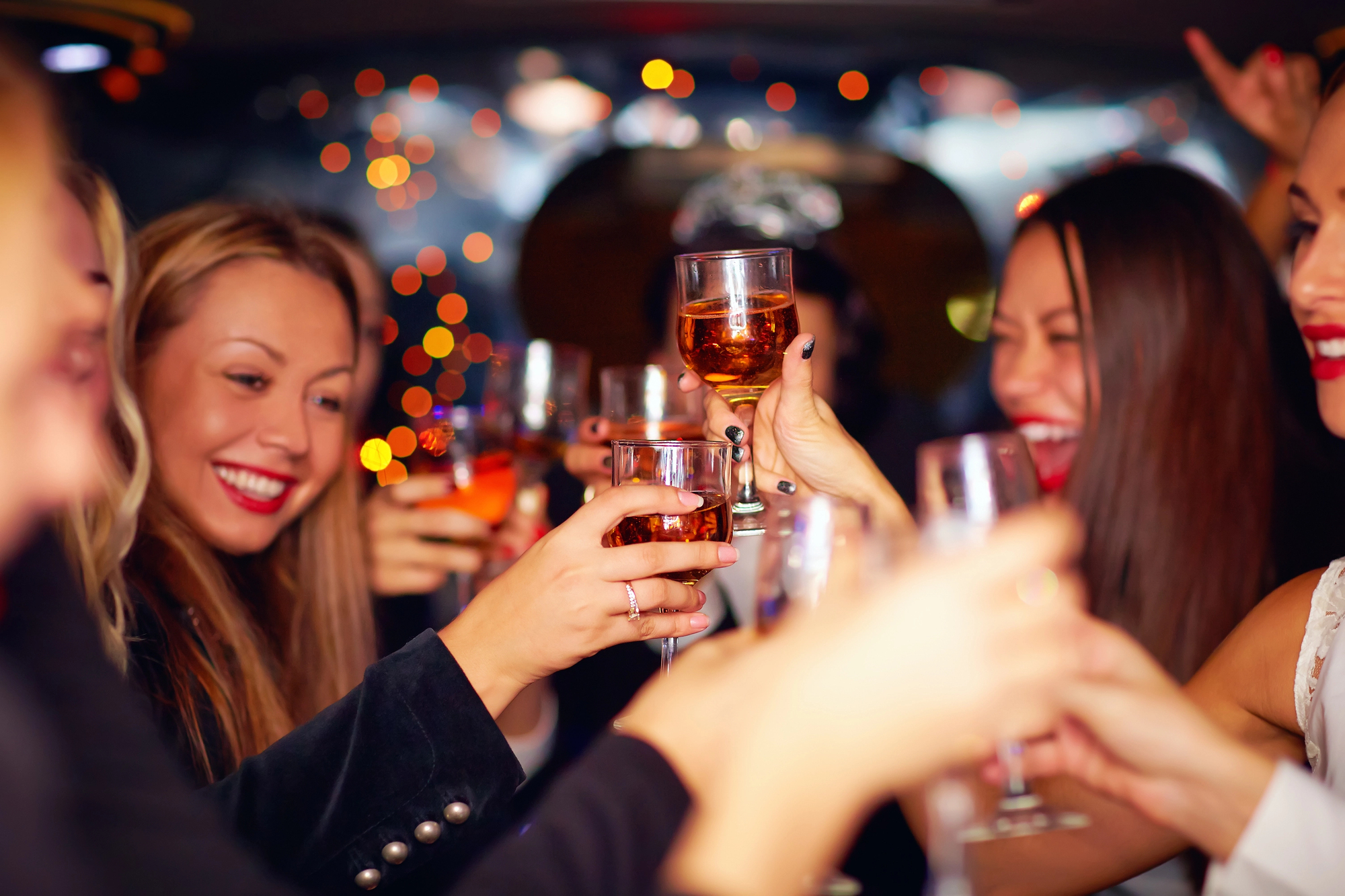 A group of smiling people raise their glasses for a toast inside a warmly lit space. Some hold champagne while others have whiskey. Festive lights in the background add to the celebratory atmosphere.