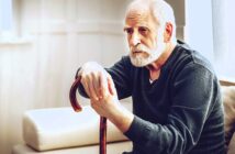 An elderly man with a white beard is sitting on a sofa, thoughtfully holding a wooden cane. He is wearing a dark sweater, and soft light filters through a nearby window, casting a gentle glow in the room.