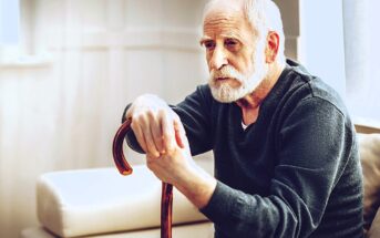 An elderly man with a white beard is sitting on a sofa, thoughtfully holding a wooden cane. He is wearing a dark sweater, and soft light filters through a nearby window, casting a gentle glow in the room.