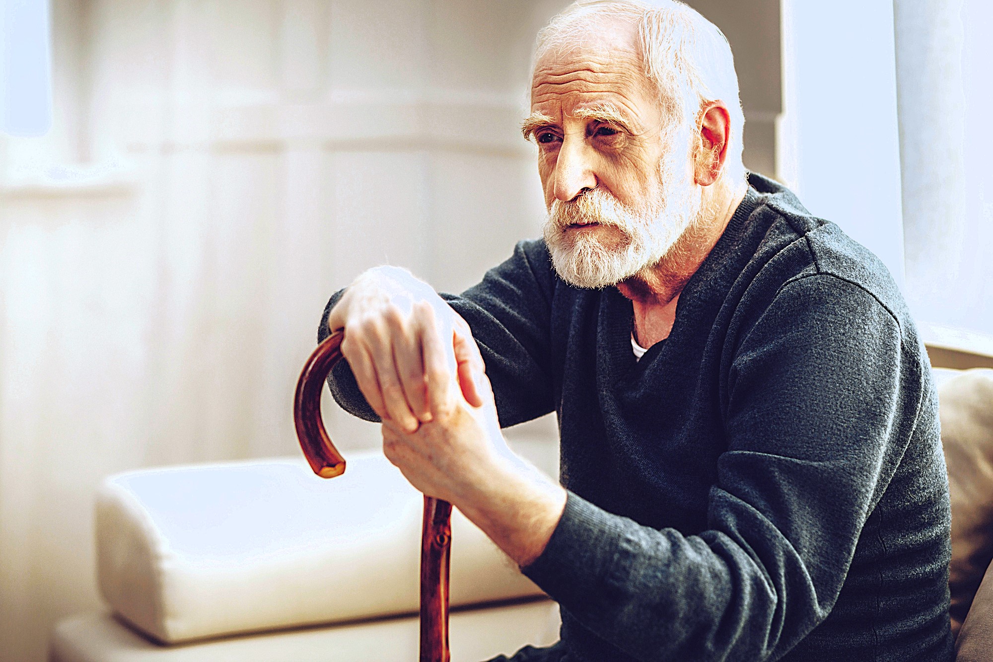 An elderly man with a white beard is sitting on a sofa, thoughtfully holding a wooden cane. He is wearing a dark sweater, and soft light filters through a nearby window, casting a gentle glow in the room.