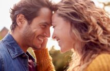 A smiling couple stands face-to-face, leaning their foreheads together. The man is wearing a denim jacket, and the woman is in a yellow sweater. They are outdoors with a blurred background, suggesting a sunny day.