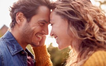 A smiling couple stands face-to-face, leaning their foreheads together. The man is wearing a denim jacket, and the woman is in a yellow sweater. They are outdoors with a blurred background, suggesting a sunny day.