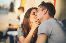 A young couple sitting closely together outdoors, sharing an intimate moment. The woman gently touches the man's chin, both smiling softly. Sunlight highlights their faces, creating a warm and relaxed atmosphere.