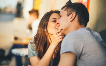 A young couple sitting closely together outdoors, sharing an intimate moment. The woman gently touches the man's chin, both smiling softly. Sunlight highlights their faces, creating a warm and relaxed atmosphere.