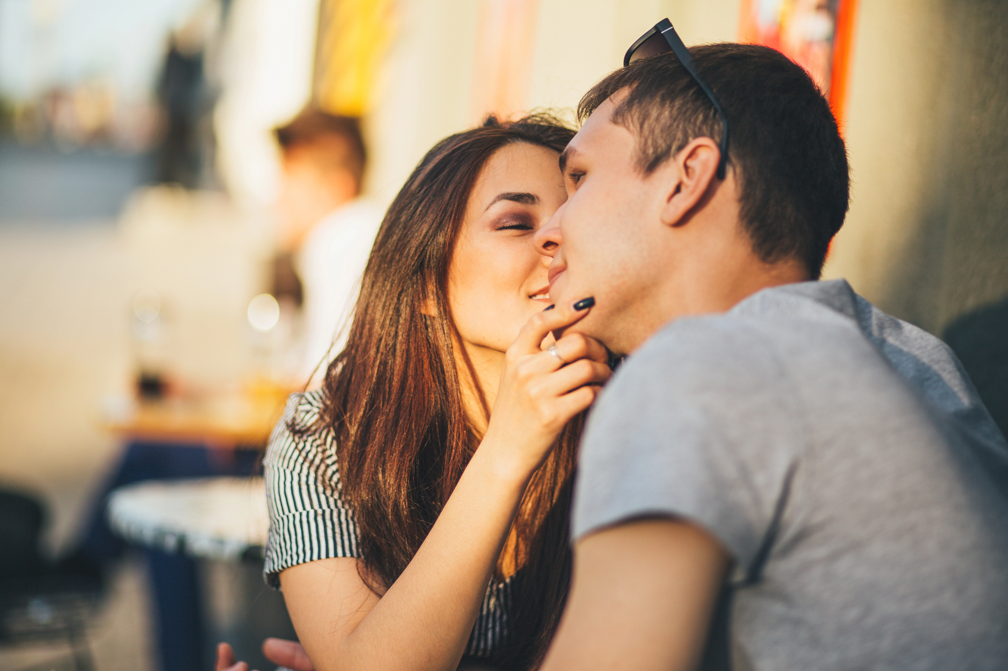 A young couple sitting closely together outdoors, sharing an intimate moment. The woman gently touches the man's chin, both smiling softly. Sunlight highlights their faces, creating a warm and relaxed atmosphere.