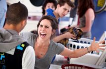 A woman is angrily yelling at a man in a laundromat. She has brown hair and is wearing a gray shirt while raising her arms. The man is facing away, wearing a black jacket. Other people in the background watch the confrontation.