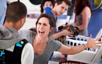 A woman is angrily yelling at a man in a laundromat. She has brown hair and is wearing a gray shirt while raising her arms. The man is facing away, wearing a black jacket. Other people in the background watch the confrontation.