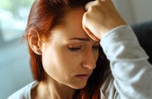 A woman with red hair is sitting, appearing upset. Her hand is resting on her forehead, and a tear is visible on her cheek. She is wearing earrings and a light-colored top. The background is softly blurred.