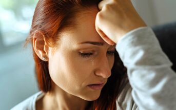 A woman with red hair is sitting, appearing upset. Her hand is resting on her forehead, and a tear is visible on her cheek. She is wearing earrings and a light-colored top. The background is softly blurred.