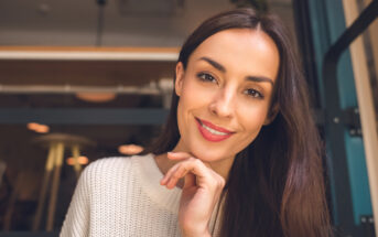 A woman with long brown hair wearing a white sweater smiles at the camera, resting her chin on her hand. She is sitting indoors with a blurred background featuring warm lighting.