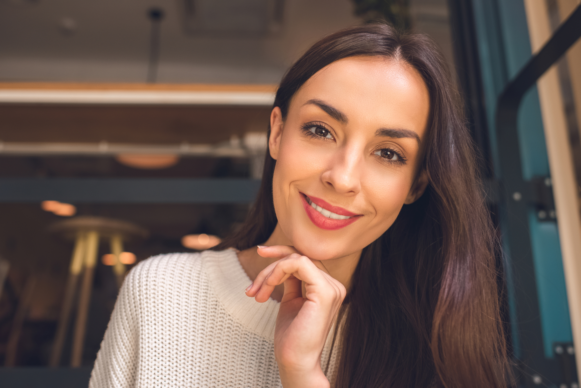 A woman with long brown hair wearing a white sweater smiles at the camera, resting her chin on her hand. She is sitting indoors with a blurred background featuring warm lighting.