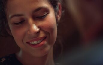 A person with curly hair and a nose ring is smiling gently, looking downward. Soft lighting creates a warm ambiance, highlighting their relaxed and contented expression.